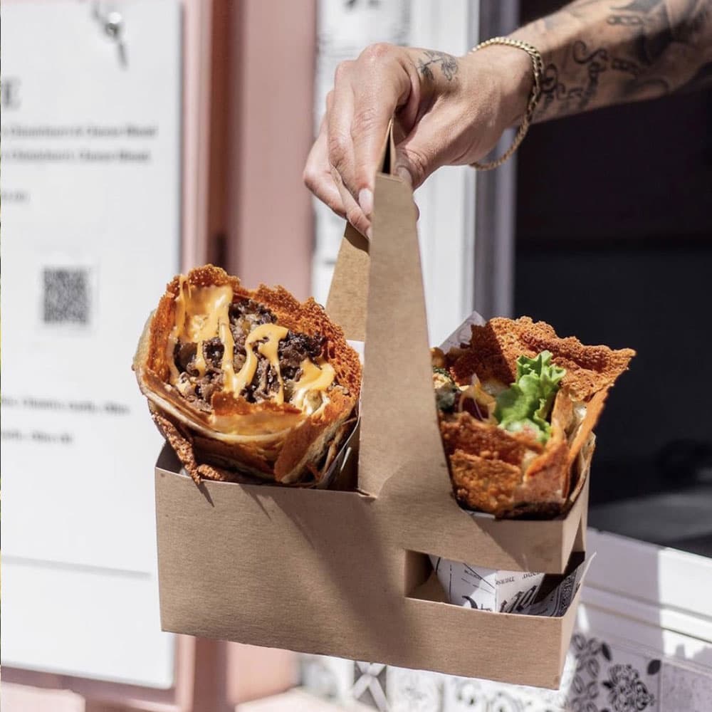 A hand holds a cardboard container with two taco-like dishes filled with ingredients.