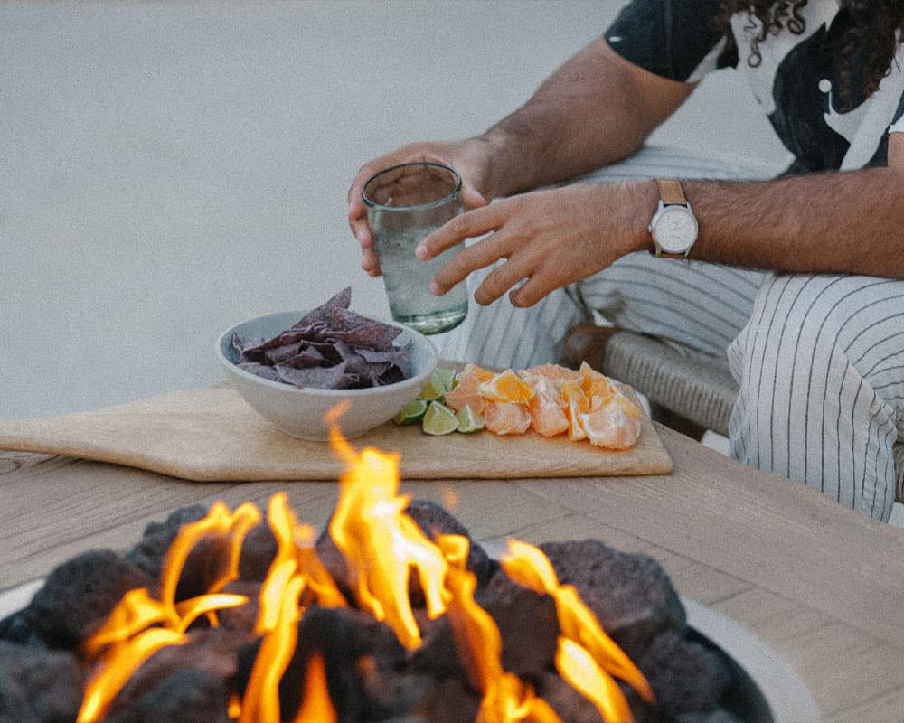 A person holding a glass sits by a fire, with a bowl of purple chips and a platter of citrus fruits nearby.