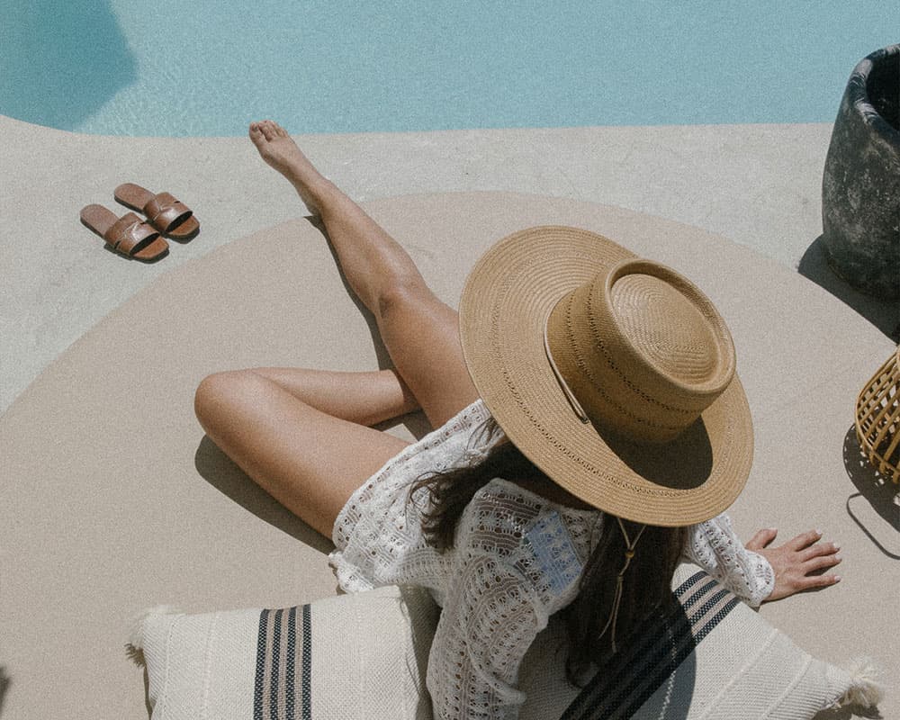 A person in a wide-brimmed hat relaxes by a poolside, surrounded by cushions and sandals.