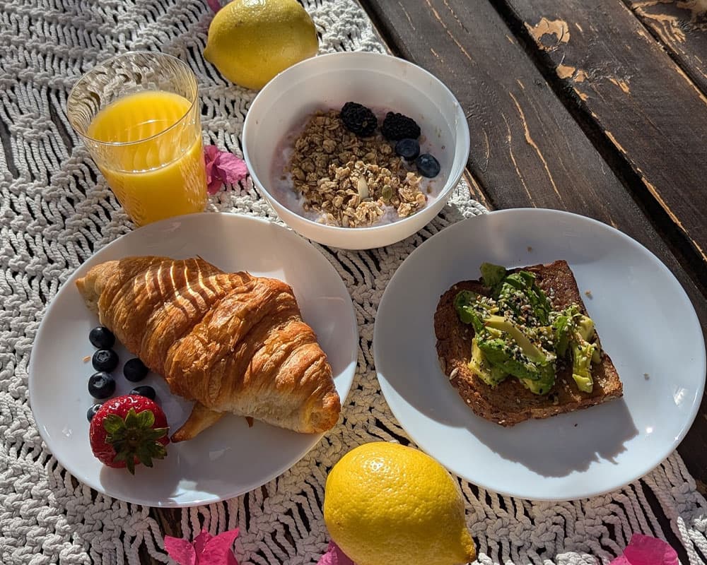 A vibrant breakfast spread featuring a croissant, avocado toast, a bowl of granola and yogurt, fresh fruit, and a glass of orange juice, all set on a rustic table.
