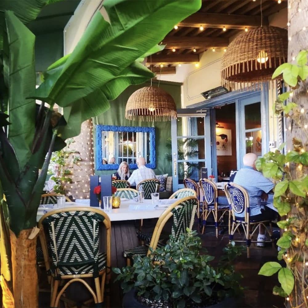 A cozy restaurant interior featuring tropical plants, woven light fixtures, and patrons seated at tables.