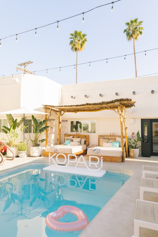 A tranquil outdoor pool area with lounge beds, palm trees, and a floating sign that reads "KOPARI."