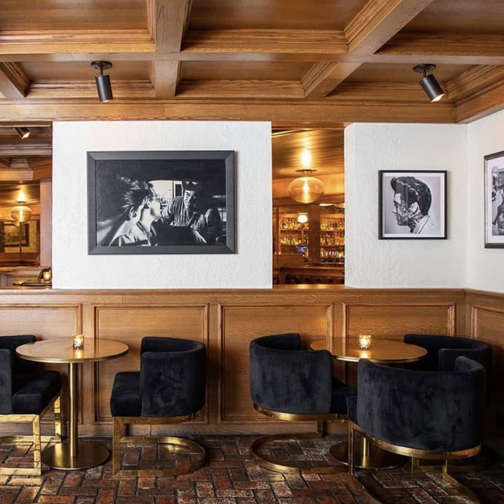 A cozy interior featuring black velvet chairs, round gold tables, and vintage black-and-white photographs on wooden walls.