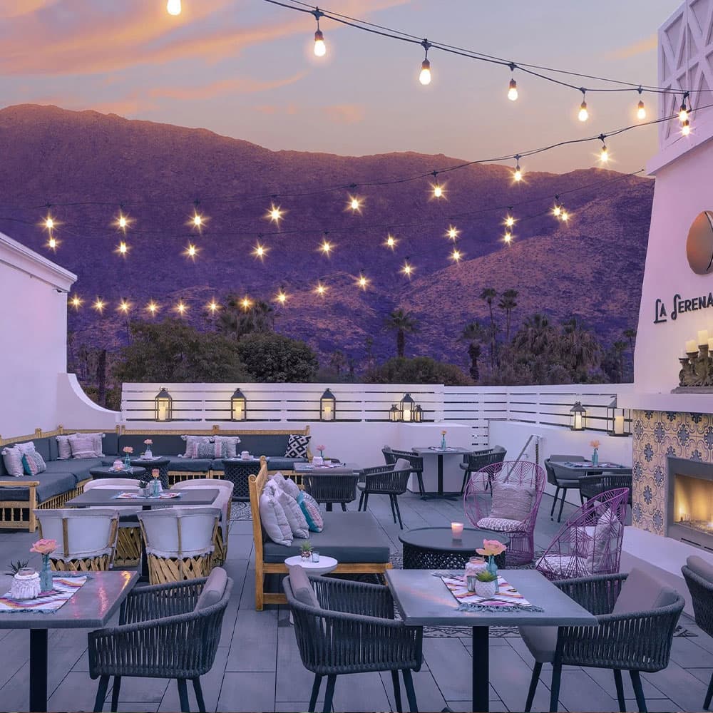 Rooftop terrace with modern seating, potted flowers, and string lights against a mountain sunset backdrop.