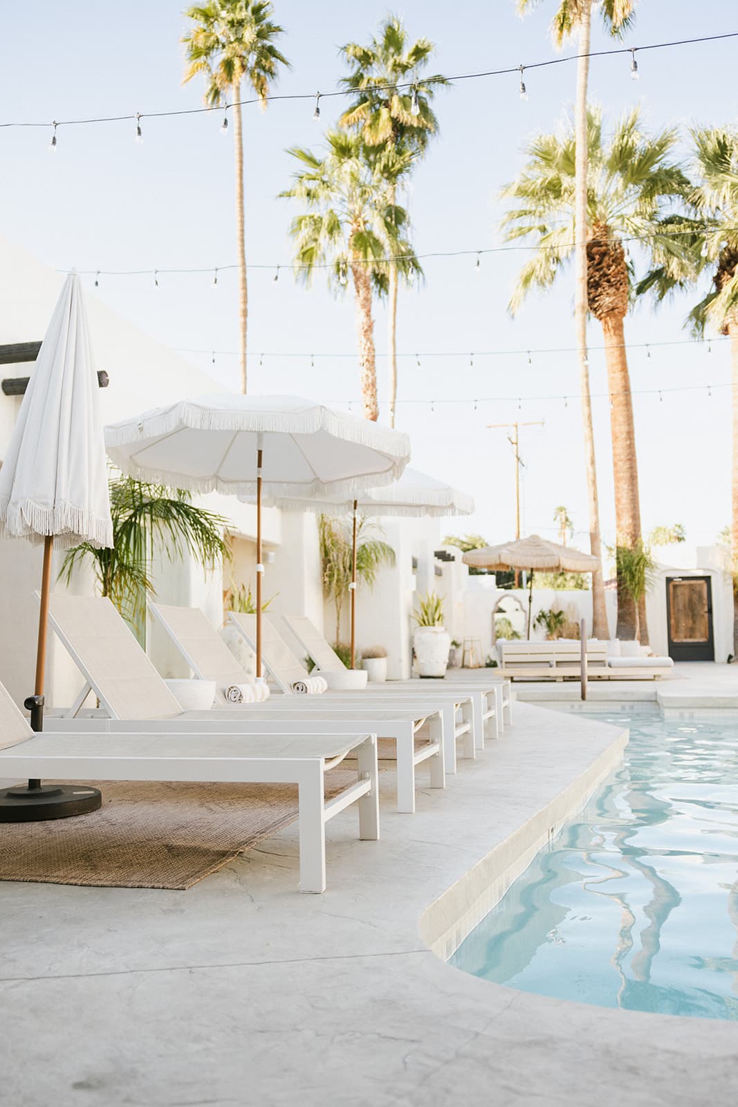 A serene pool area featuring white lounge chairs, umbrellas, and palm trees against a clear sky.