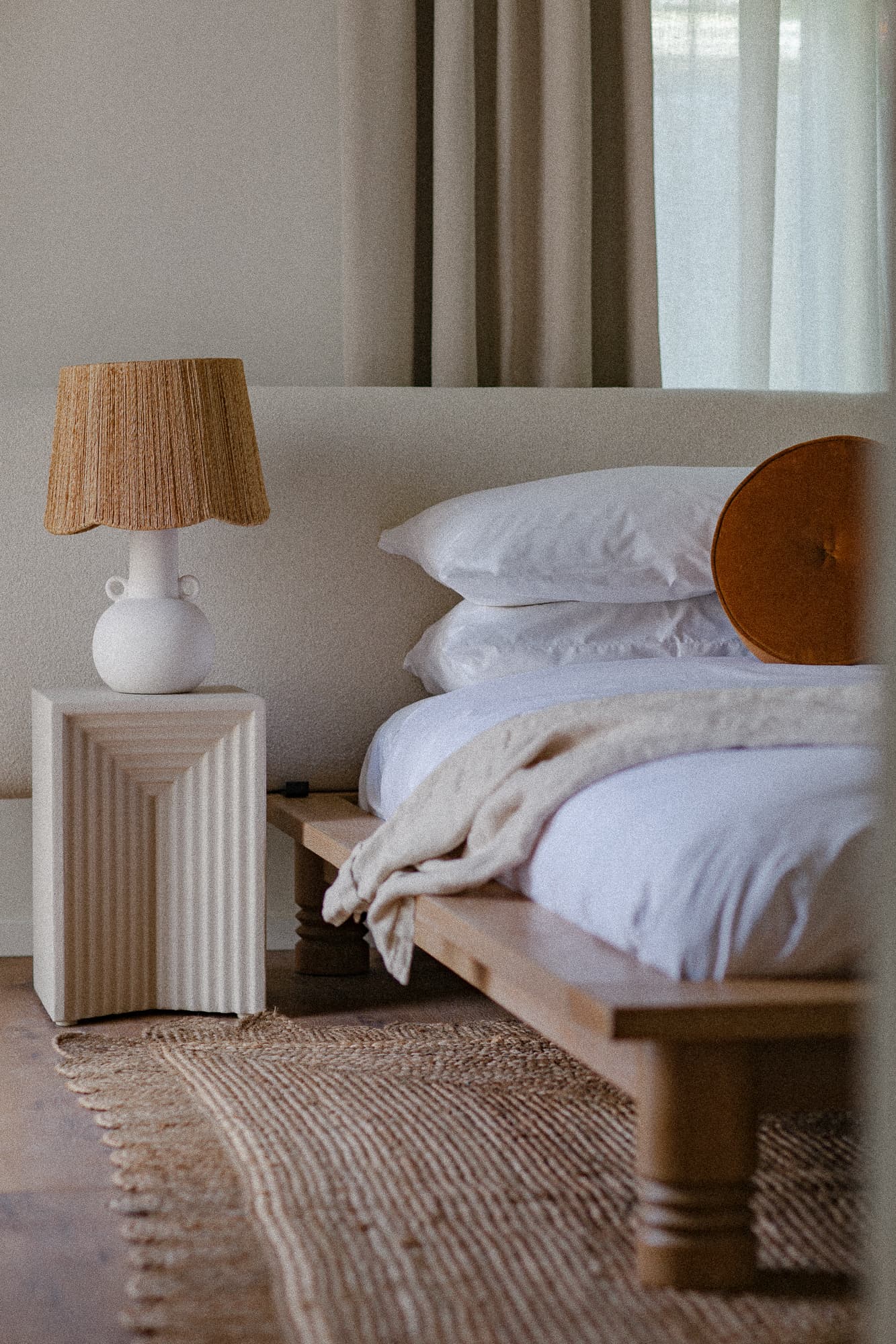 A cozy bedroom featuring a wooden bed, layered linens, a textured rug, and a unique lamp on a bedside table.